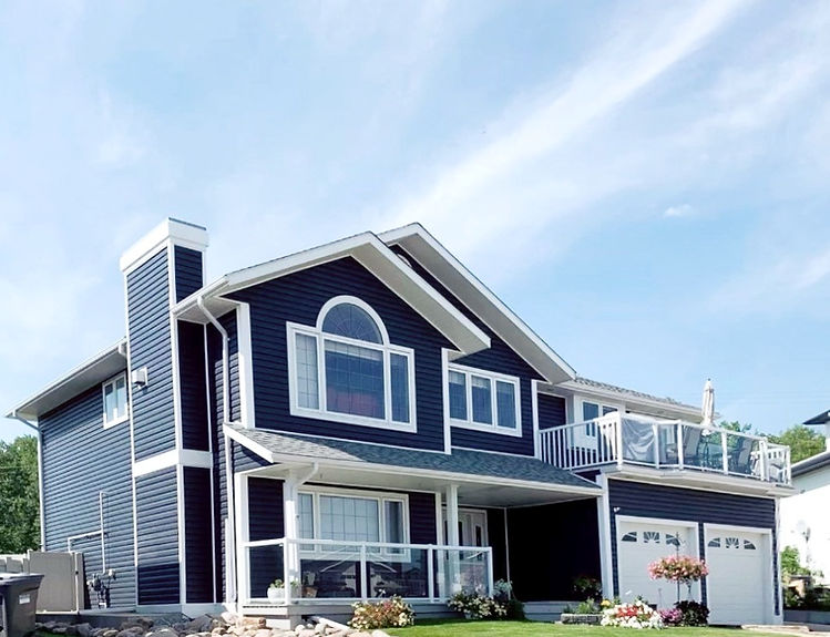 A large house with blue siding and white trim, surrounded by green trees and a clear blue sky.