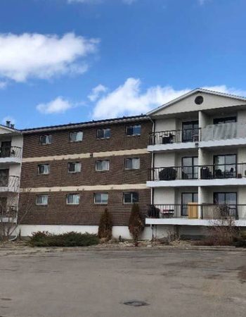 An apartment building with a parking lot in front, surrounded by trees and a clear blue sky.